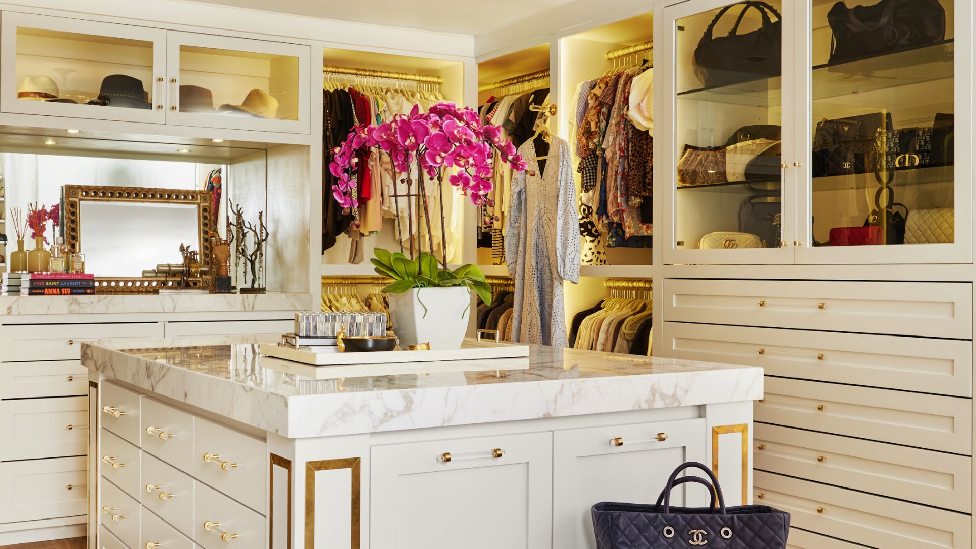 An opulent walk-in closet with white cabinetry, gold hardware, and marble island topped with vibrant orchids.
