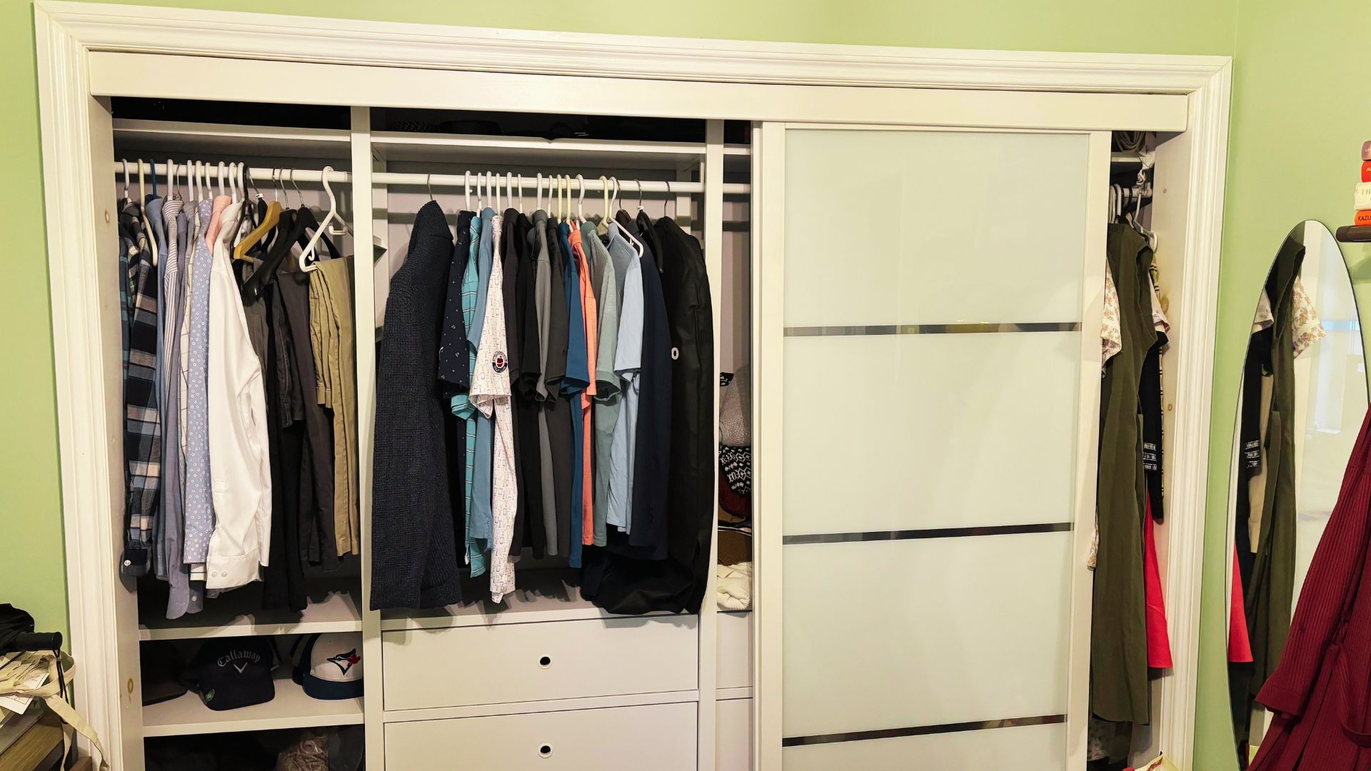 White sliding-door wardrobe with one door open, revealing neatly hung shirts, jackets, and polos sorted by color.