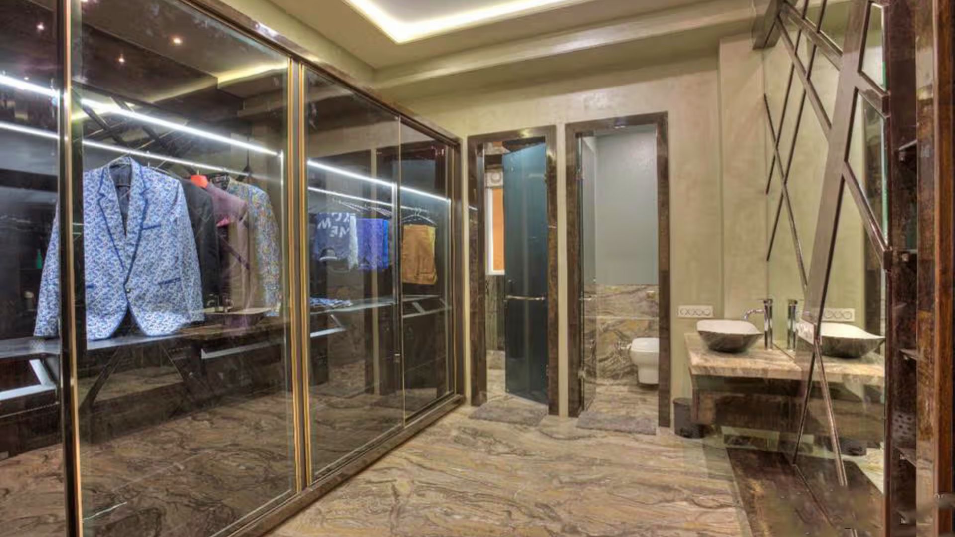 A high-end dressing suite with floor-to-ceiling glass cabinets displaying designer suits, marble floors, and integrated LED lighting.