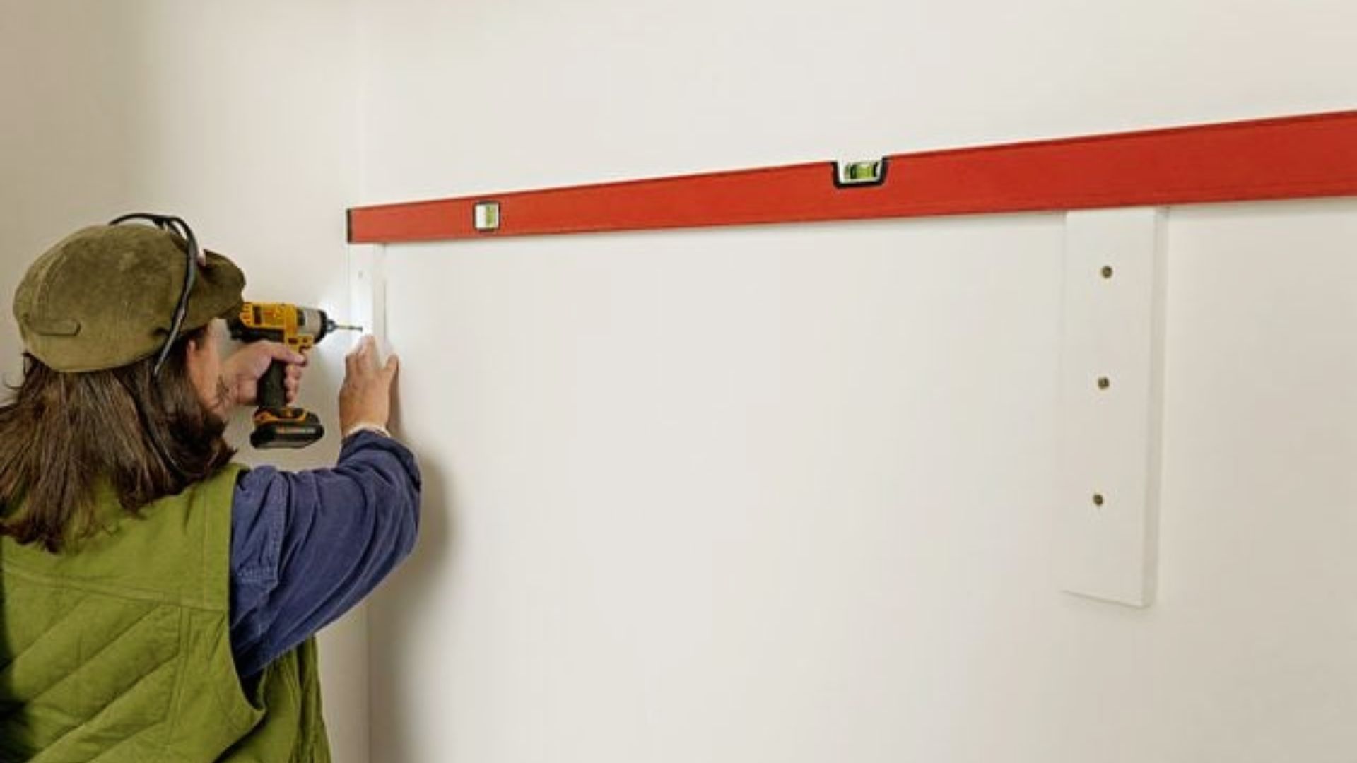 Person in a green vest and cap using a power drill to install white support boards on a closet wall with a red level above.