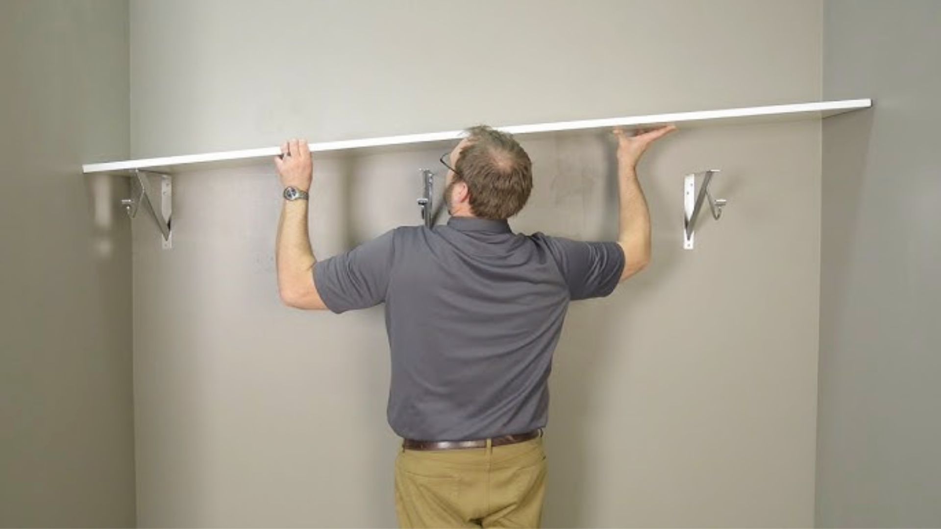 Man in gray polo shirt installing a white shelf above metal closet rod brackets on a bare wall.