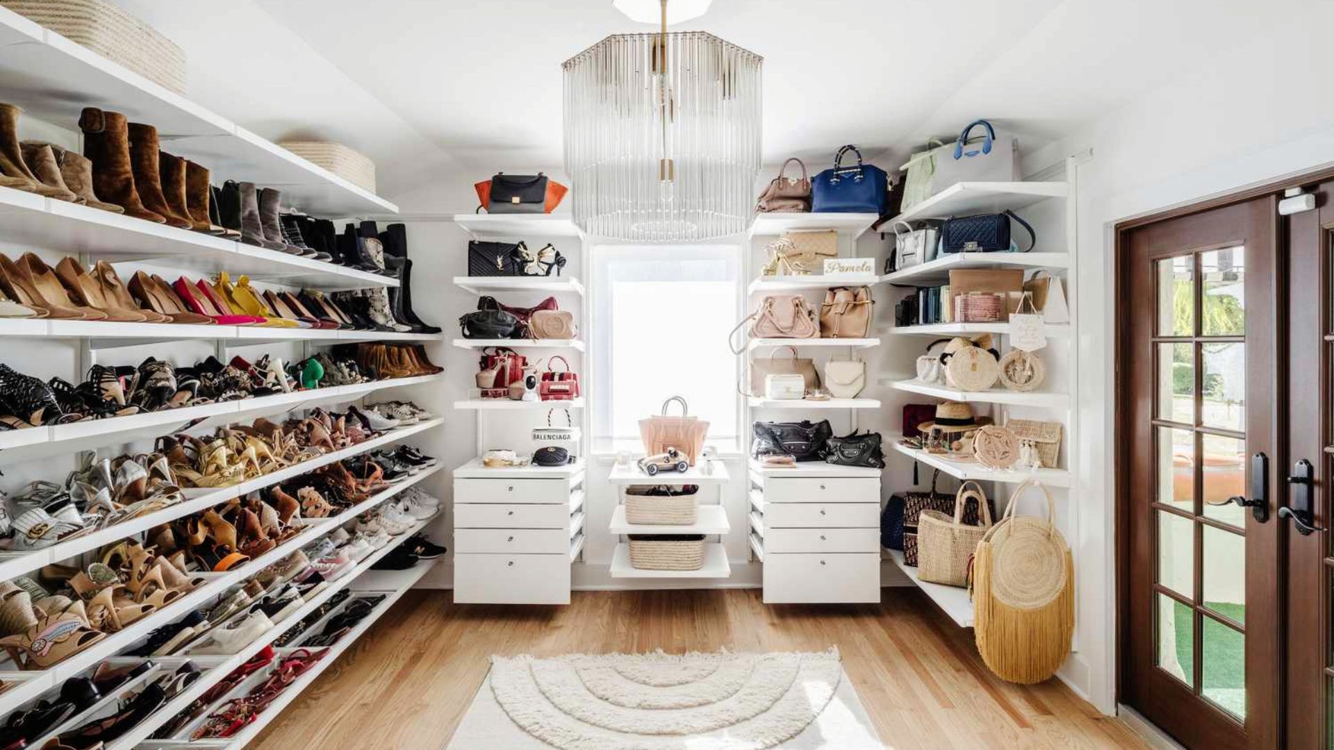A bright walk-in closet lined with floor-to-ceiling white shelves holding hundreds of designer shoes and handbags.