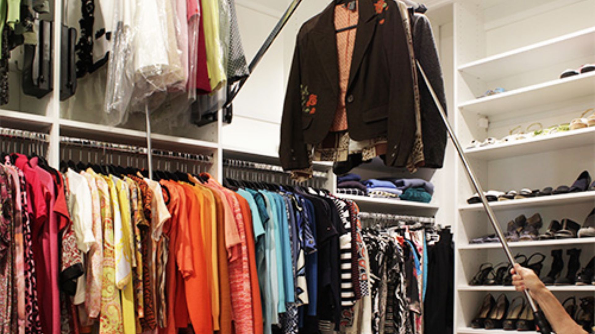 Person using a long metal pole to lower a pull-down closet rod with jackets in a large walk-in closet.
