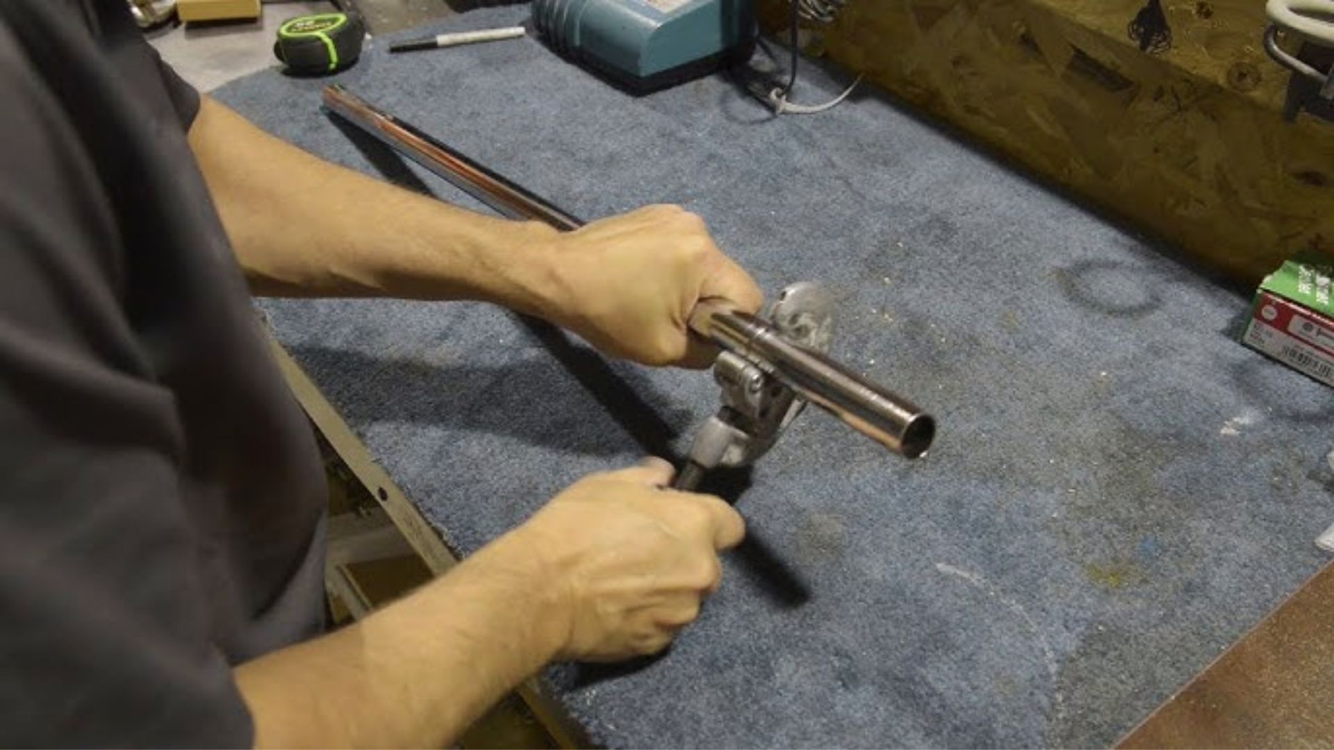 Person using a pipe cutter to trim a chrome closet rod on a blue carpeted workbench with tools nearby.