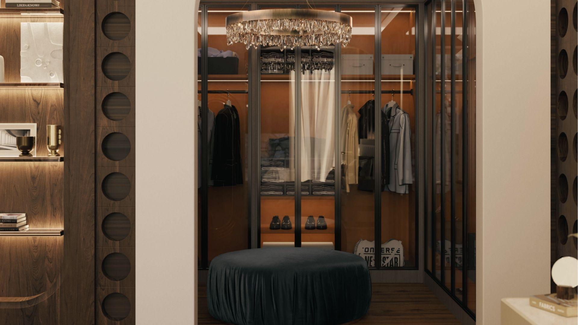 Luxe walk-in closet with glass doors, crystal chandelier, velvet ottoman, and wood shelving.